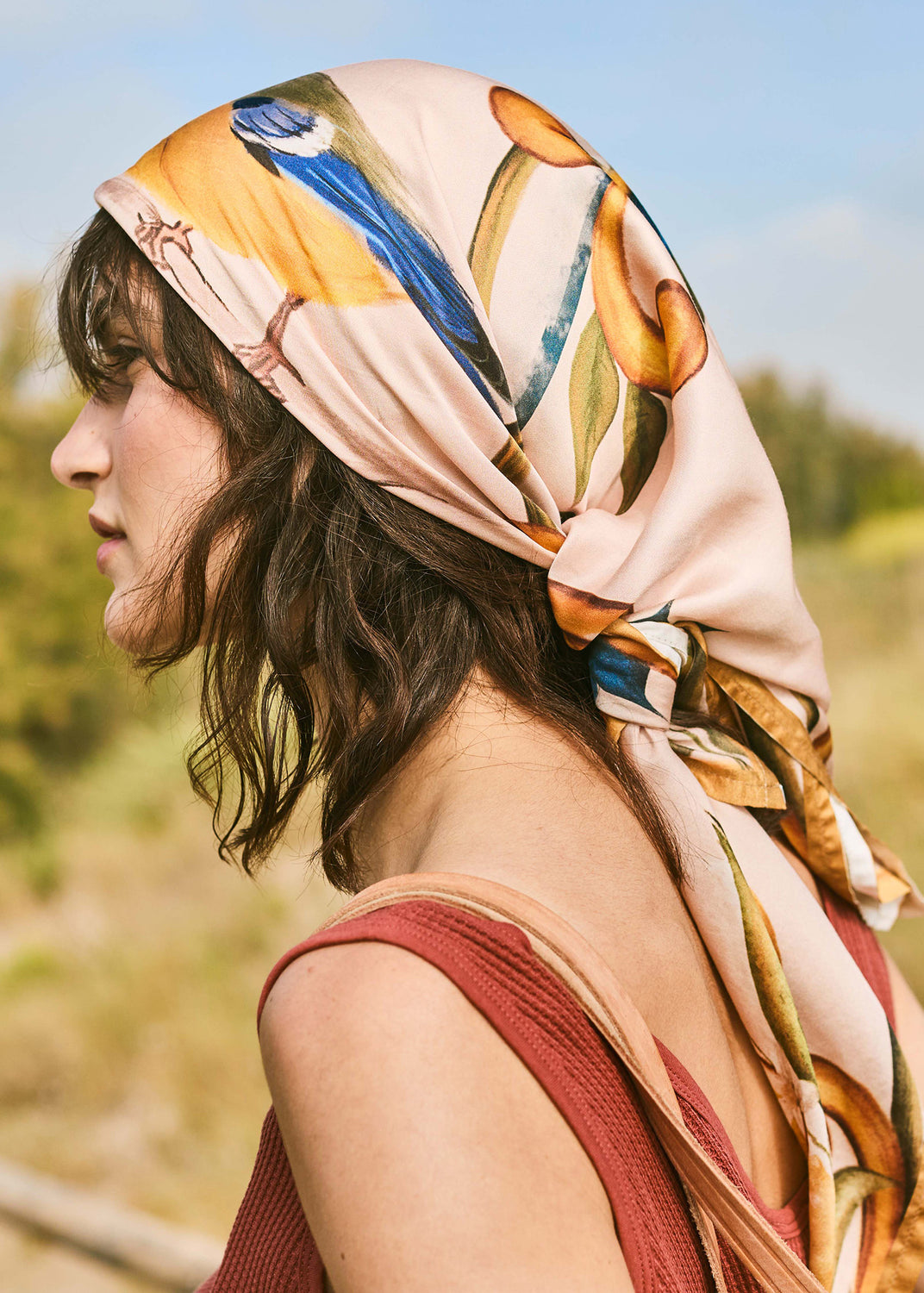 Woman wearing a robin print square scarf tied over her hair in a warm Mediterranean outdoor setting.