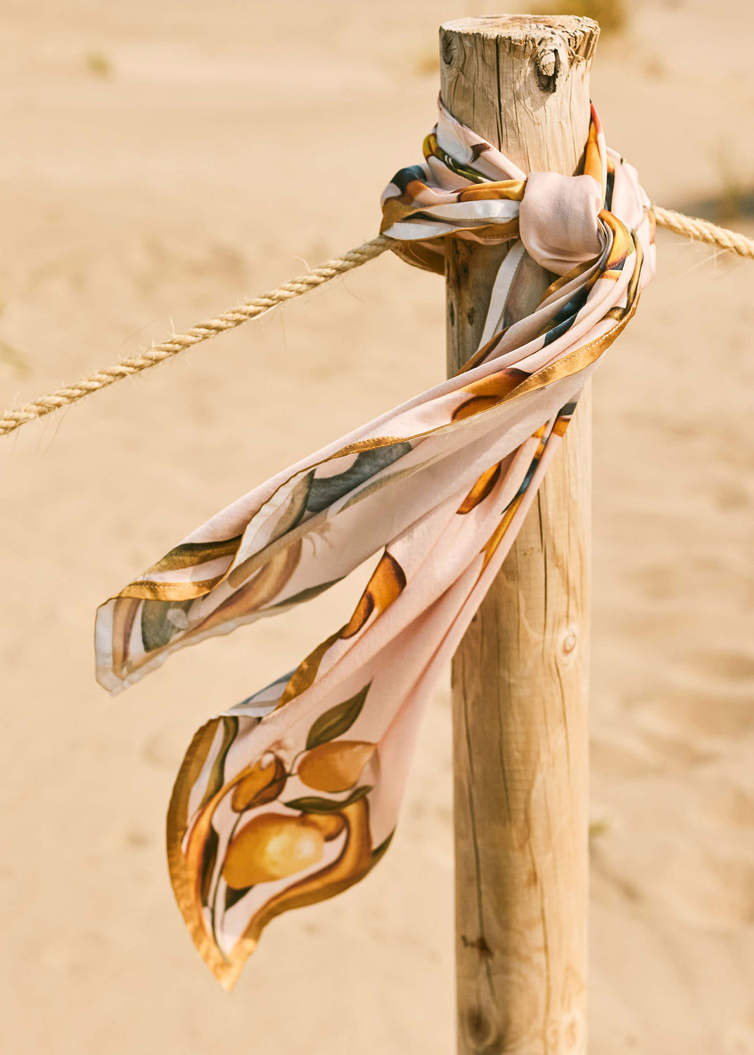 Robin print scarf tied around a wooden post on a sandy Mediterranean beach in warm natural light.