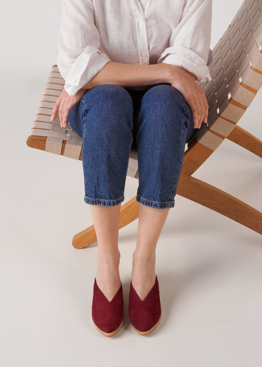 Woman seated in a woven lounge chair wearing burgundy espadrille mule wedges styled with a white linen shirt and dark denim jeans in a clean studio setting