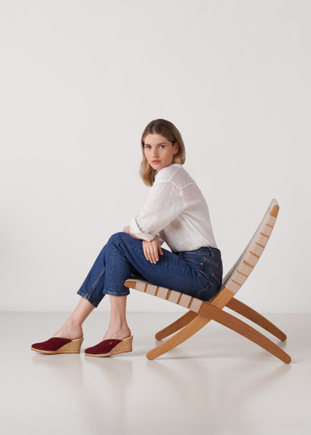 Woman seated in a woven lounge chair wearing burgundy wedge espadrille mules styled with a white linen shirt and cropped denim jeans in a clean studio setting
