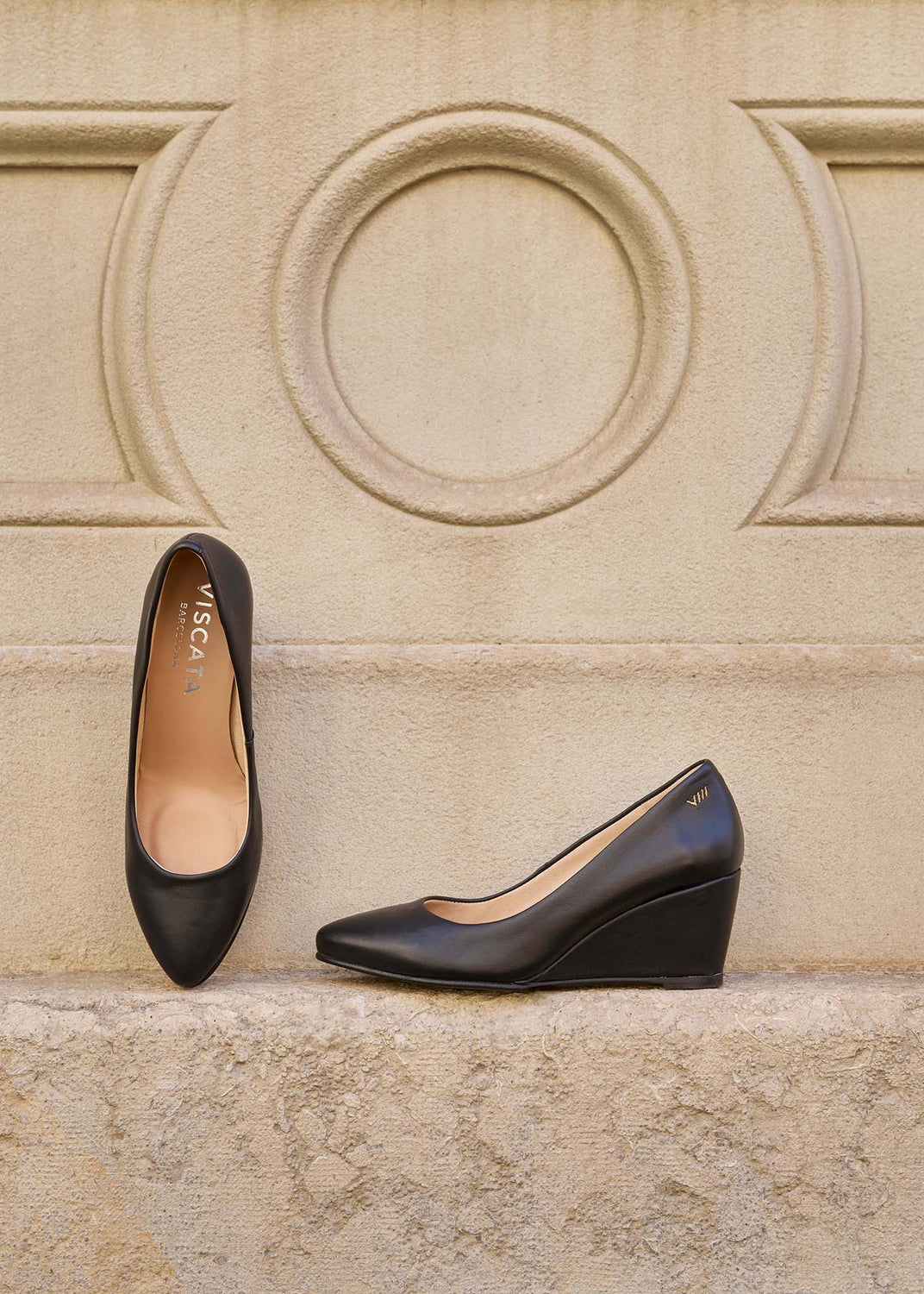 A pair of black pointed-toe wedge pumps by VISCATA, one shown from the top and one from the side, resting on a textured stone ledge against a beige stone wall.