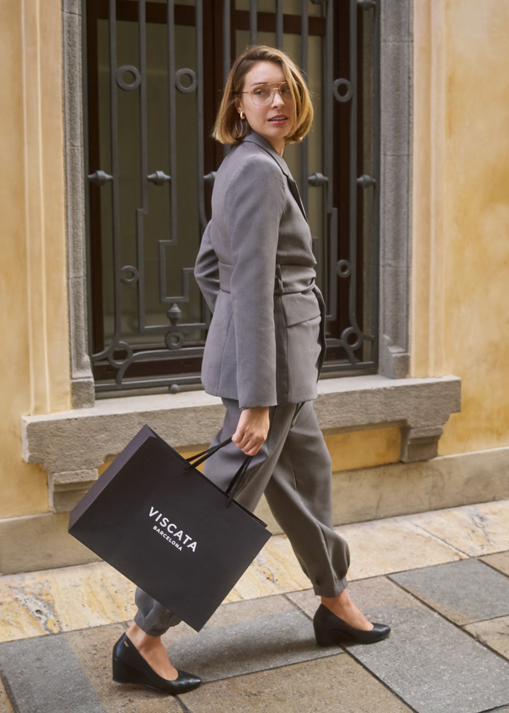 Woman walking confidently in a tailored grey suit paired with black wedge shoes, holding a Viscata Barcelona shopping bag — Viscata Essenza business chic look.