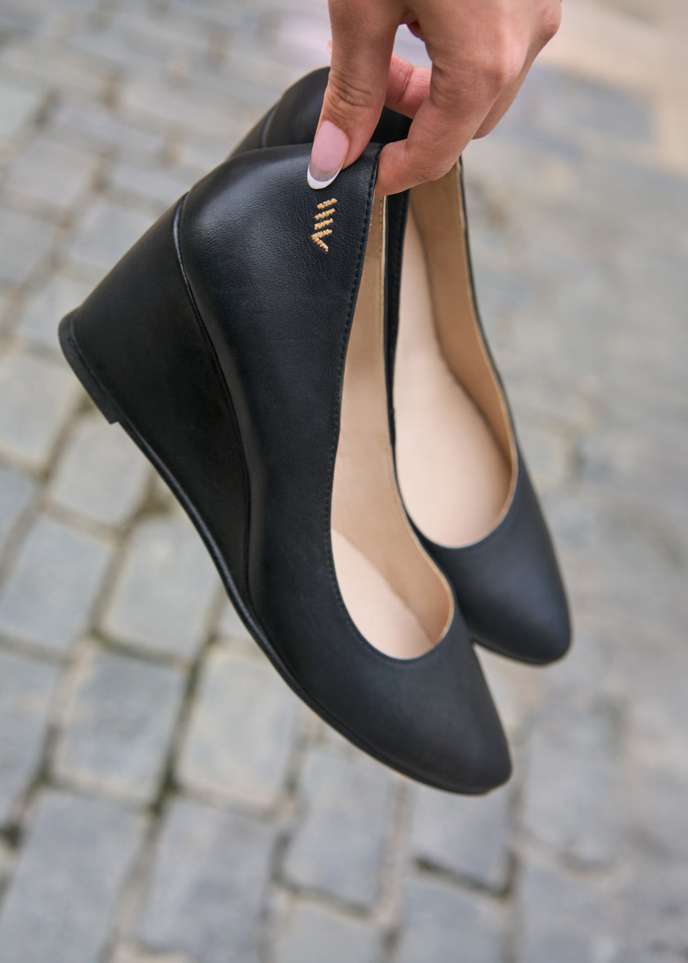 Close-up of black leather wedge shoes with gold Viscata logo detail, held by hand over cobblestone street — Viscata Essenza collection minimalist elegance.