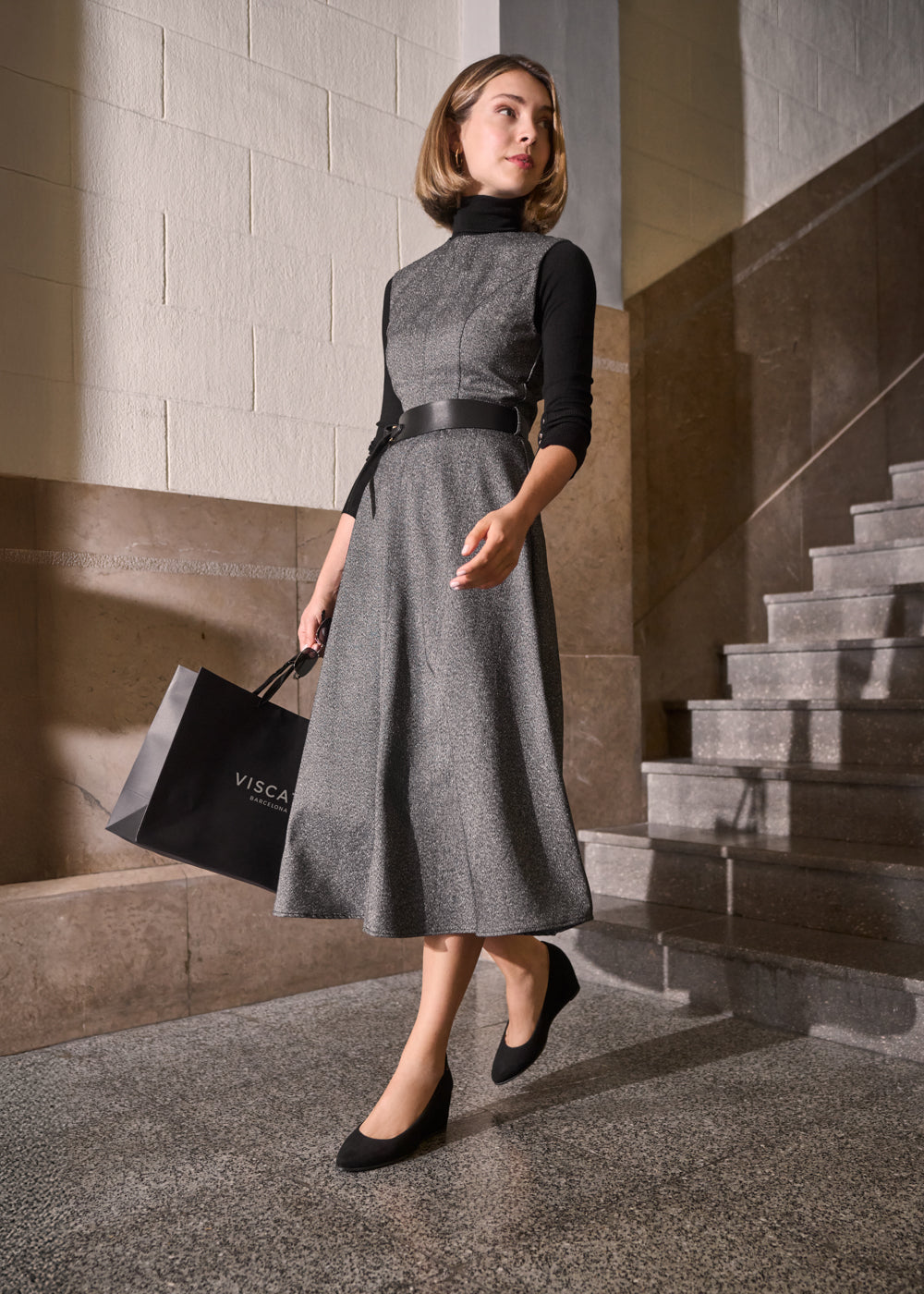 Woman walking indoors wearing a grey midi dress over a black turtleneck, styled with black wedge shoes and carrying a Viscata Barcelona bag — Essenza collection, timeless elegance in motion.
