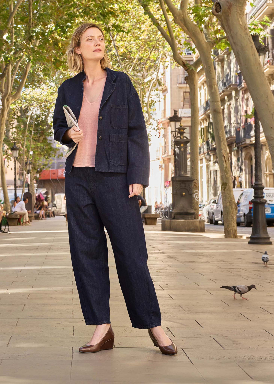 A woman stands on a sunlit city sidewalk wearing a dark navy tailored jacket and matching wide-leg trousers over a light pink top, with brown wedge shoes. Trees, buildings, and a few pigeons are visible in the background.