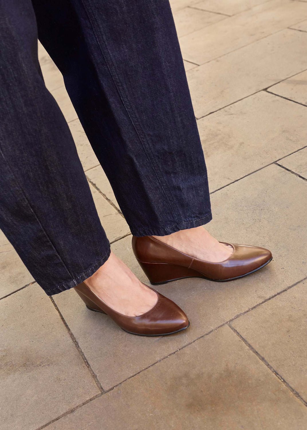 A person wearing dark brown leather wedge pumps with pointed toes, paired with dark blue jeans, standing on a light brown stone tiled floor.