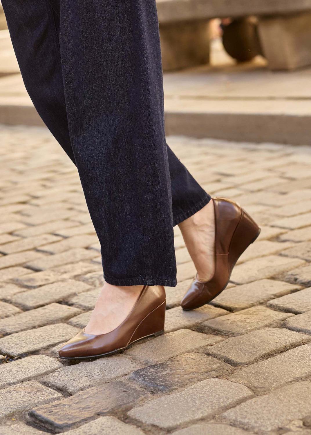 A person wearing dark navy pants and brown leather wedge pumps with pointed toes, walking on a cobblestone street.