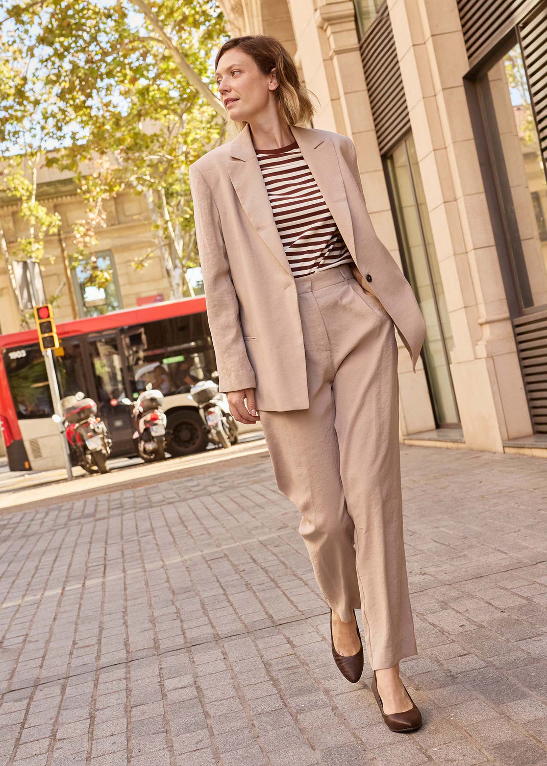 A woman walking on a city sidewalk wearing a beige tailored blazer and matching trousers over a brown and white striped top, with brown pointed flats. Trees, buildings, and a red bus are visible in the background.