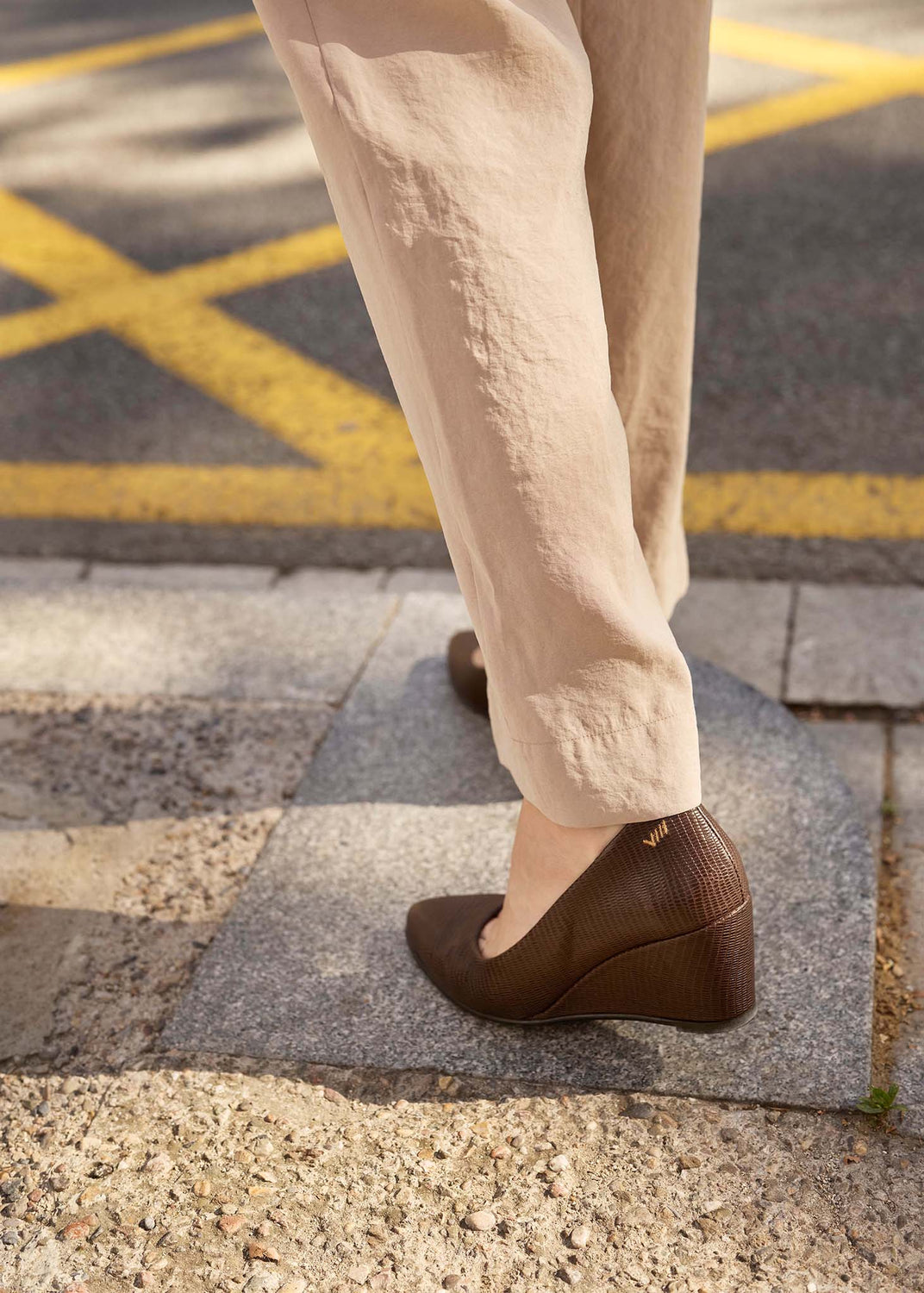 A person wearing beige trousers and brown wedge shoes stands on a stone curb outdoors near a road with yellow painted markings.