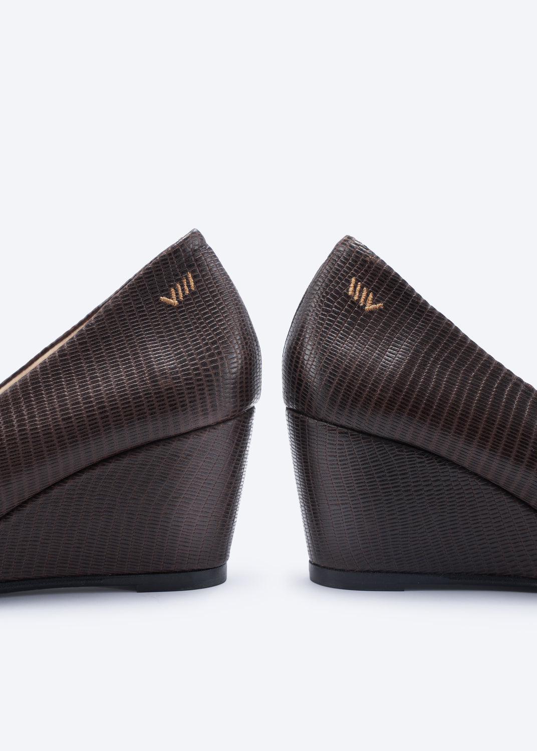 Close-up rear view of a pair of dark brown wedge pumps with an embossed reptile pattern and a small gold logo on the heel counter, set against a plain white background.