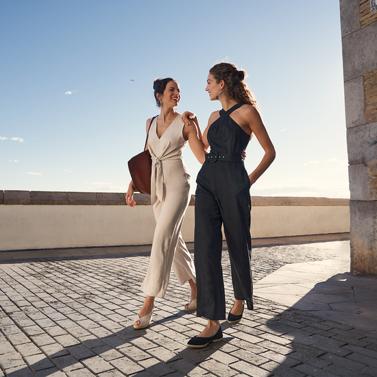 Two women walking by the sun, one wearing a beige jumpsuit paired with Verneda canvas beige slingback open toe espadrilles and the other wearing a black jumsuit paired with Garvet black suede foldable espadrille flats