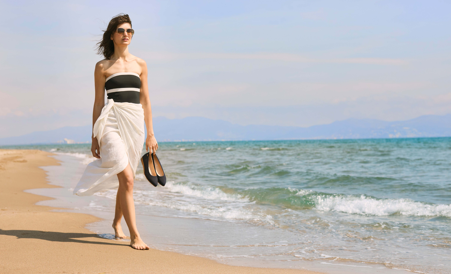 Woman in a black and white dress walking on a beach with ocean and sky in the background
