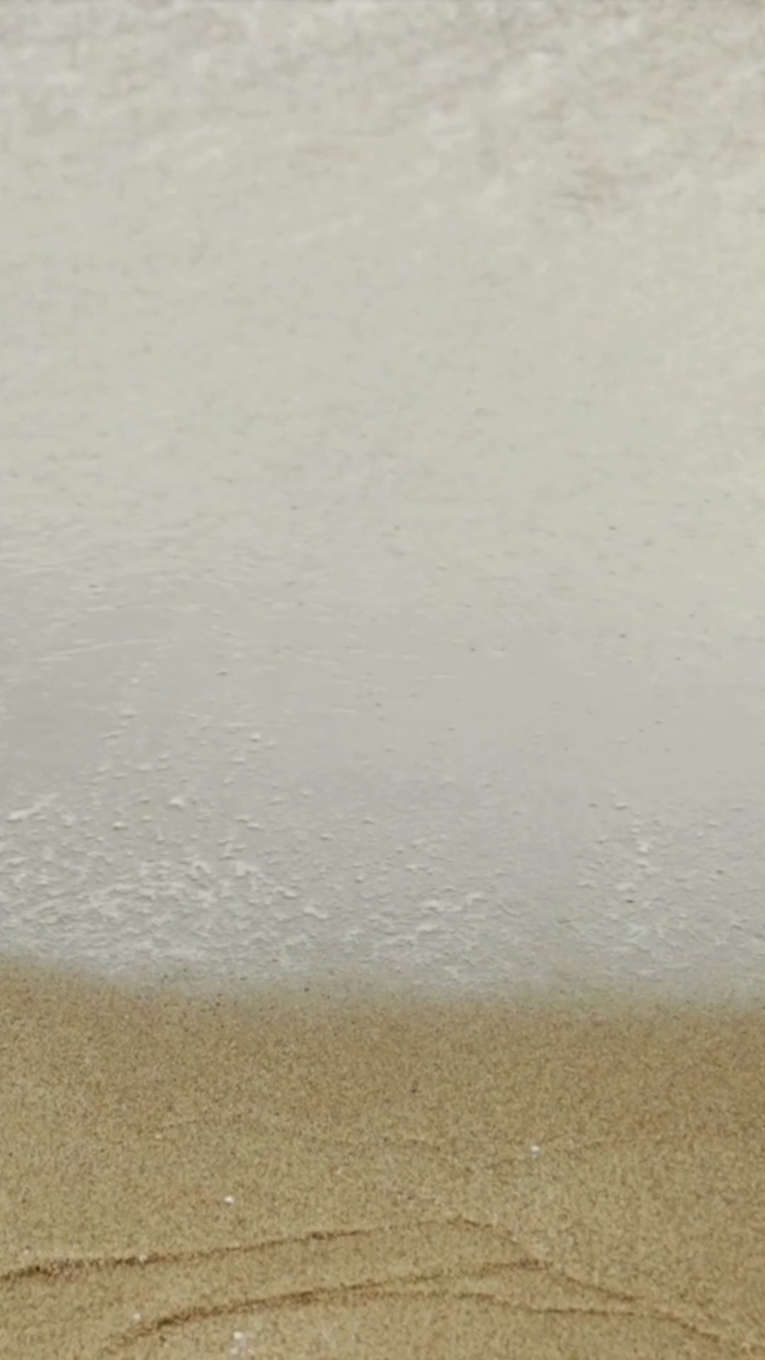 Gentle ocean waves roll onto a sandy beach, with white foam washing in and receding in a steady rhythm.